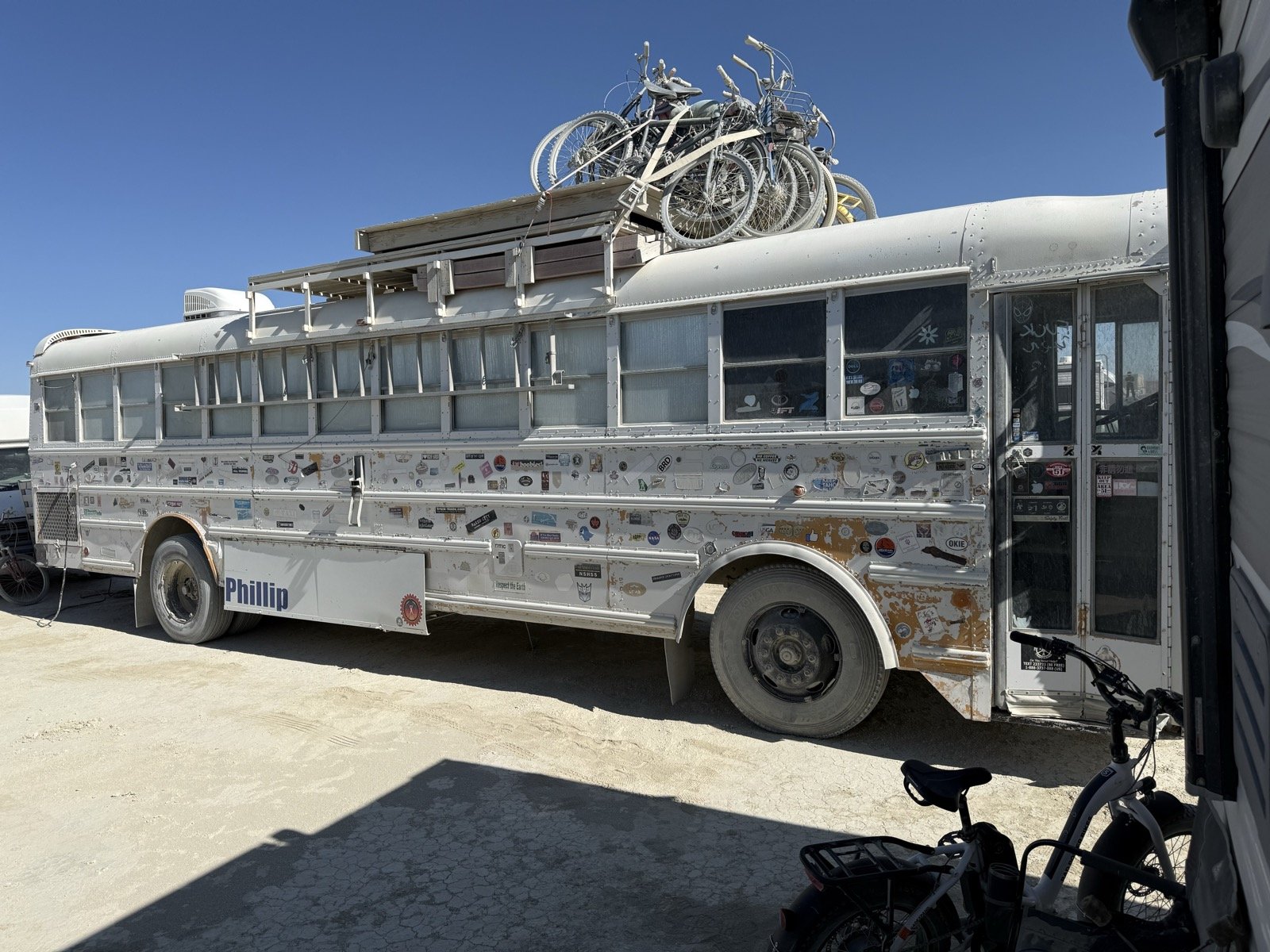 The real Phillip: a white school bus covered in stickers with bicycles strapped to a roof deck, parked on cracked playa